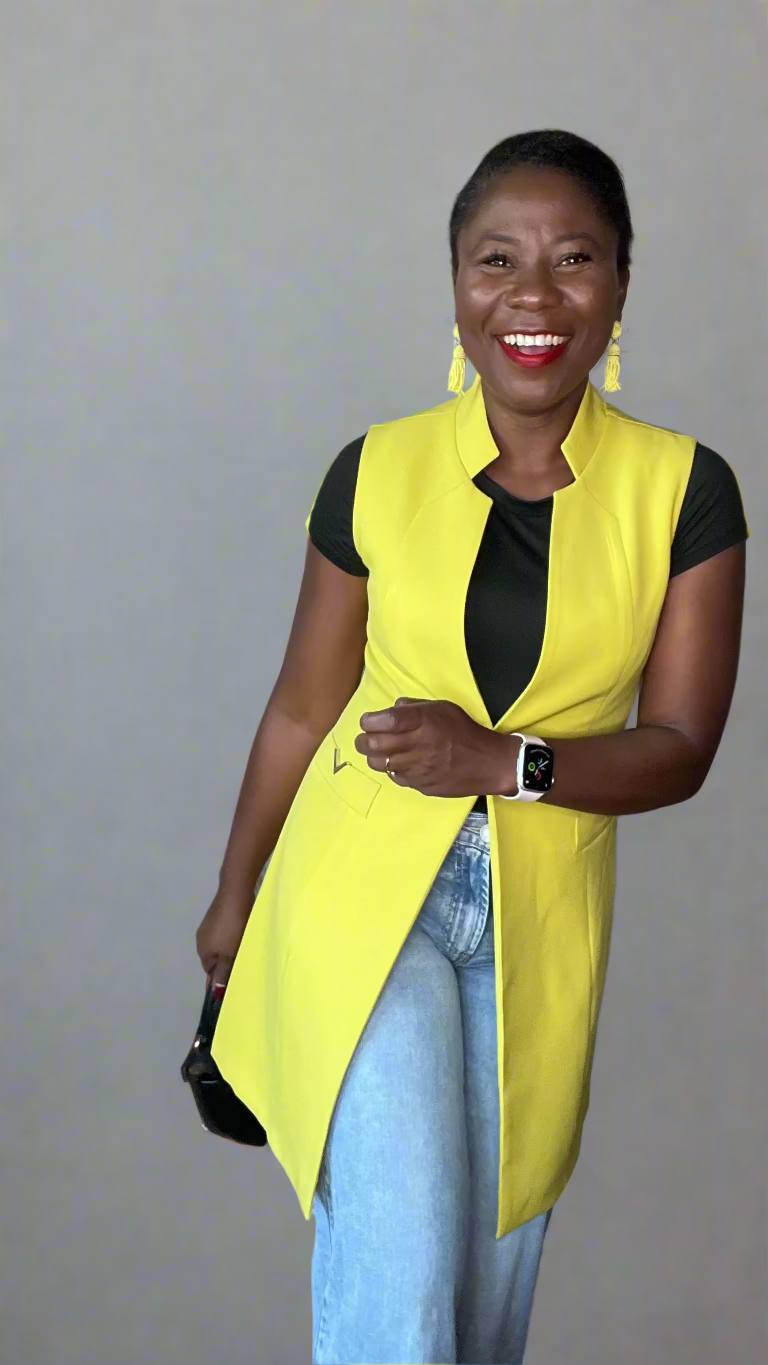 Woman wearing a bright yellow vest over a black top and light blue jeans, standing against a plain wall.