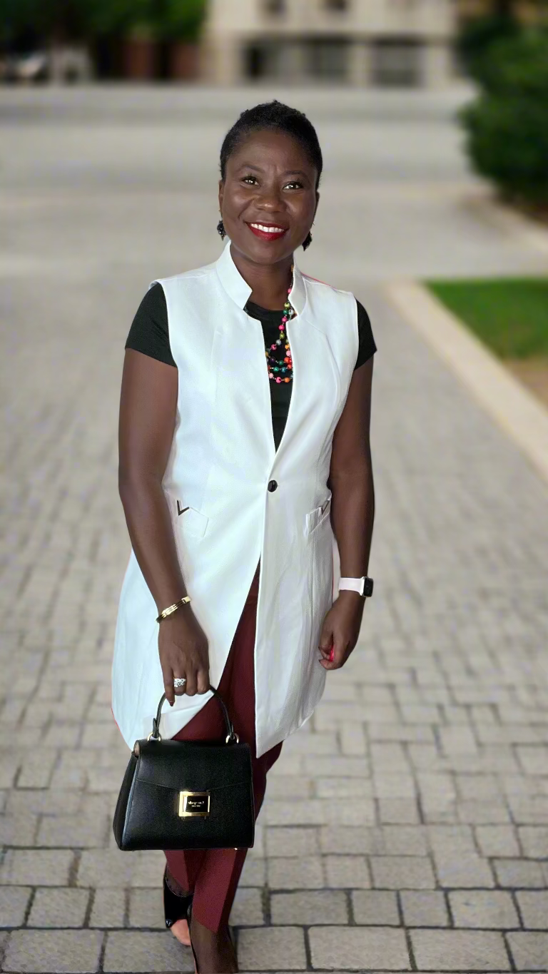Woman wearing a white vest, black top, red pants, and holding a black clutch against a plain wall.
